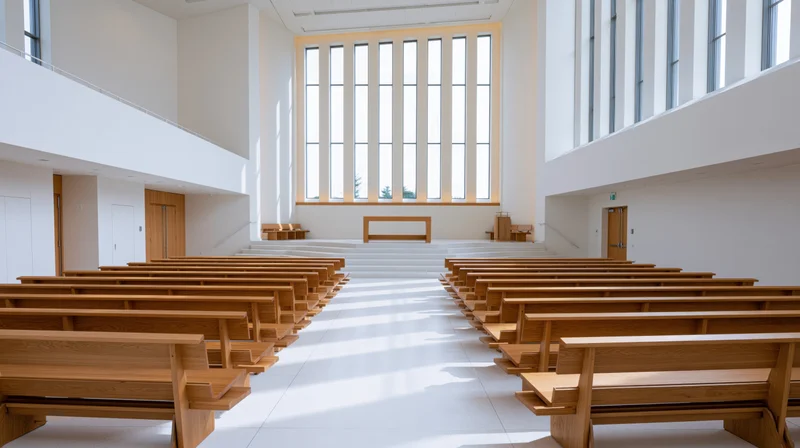 Empty wooden pews arranged in rows inside a minimalist modern church nave with white walls and large windows letting in natural light
