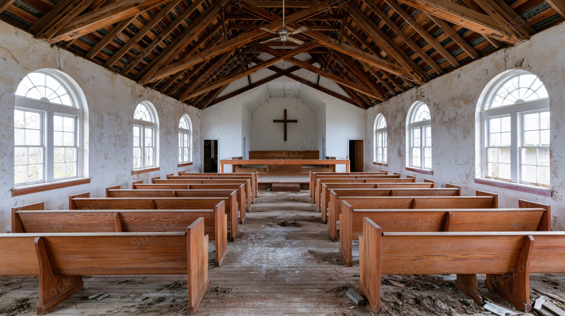 Rows of wooden pews arranged in an abandoned rural church interior with a cross on the wall and arched windows letting in natural light