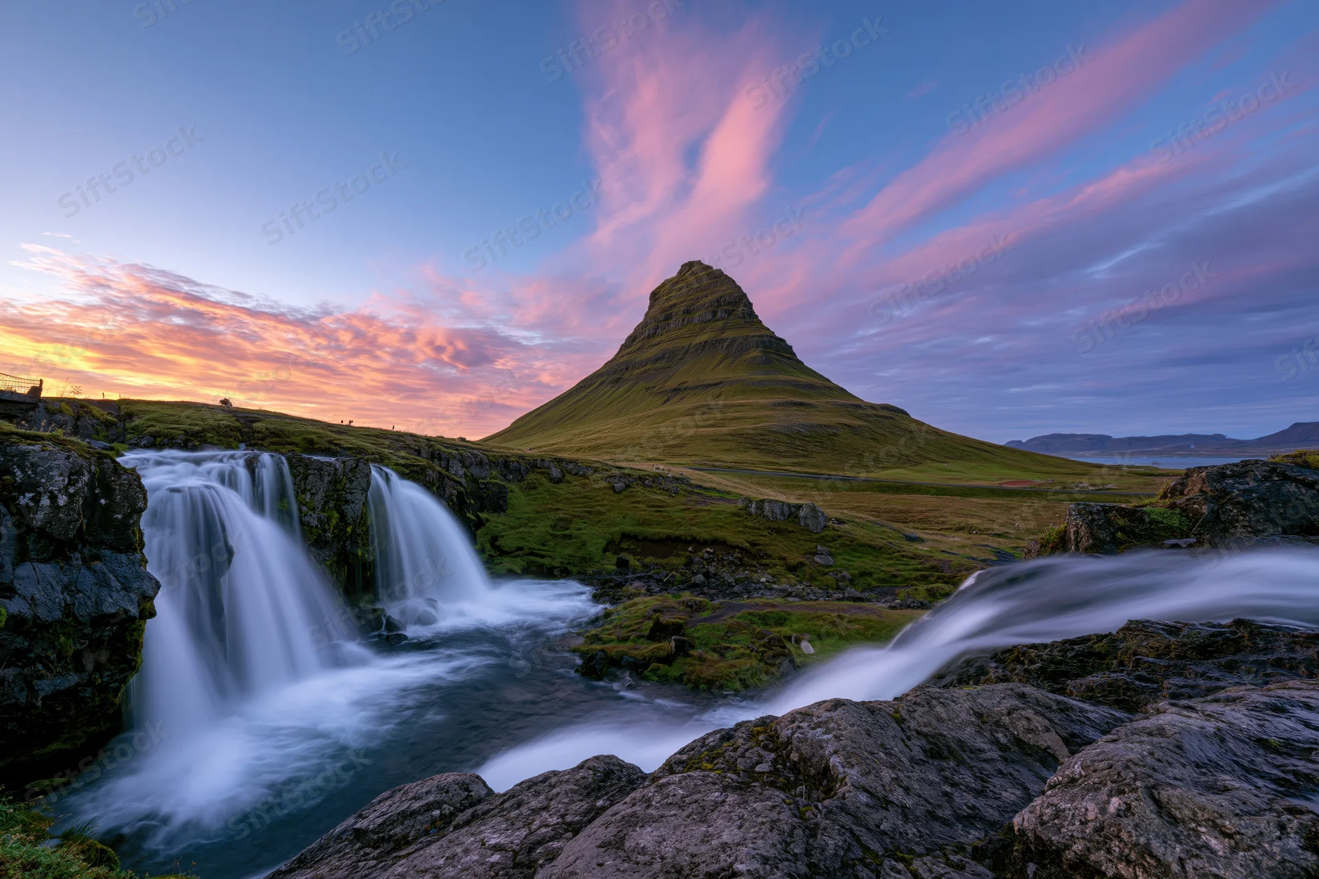 Kirkjufell mountain rising above cascading waterfalls in a grassy valley at sunset