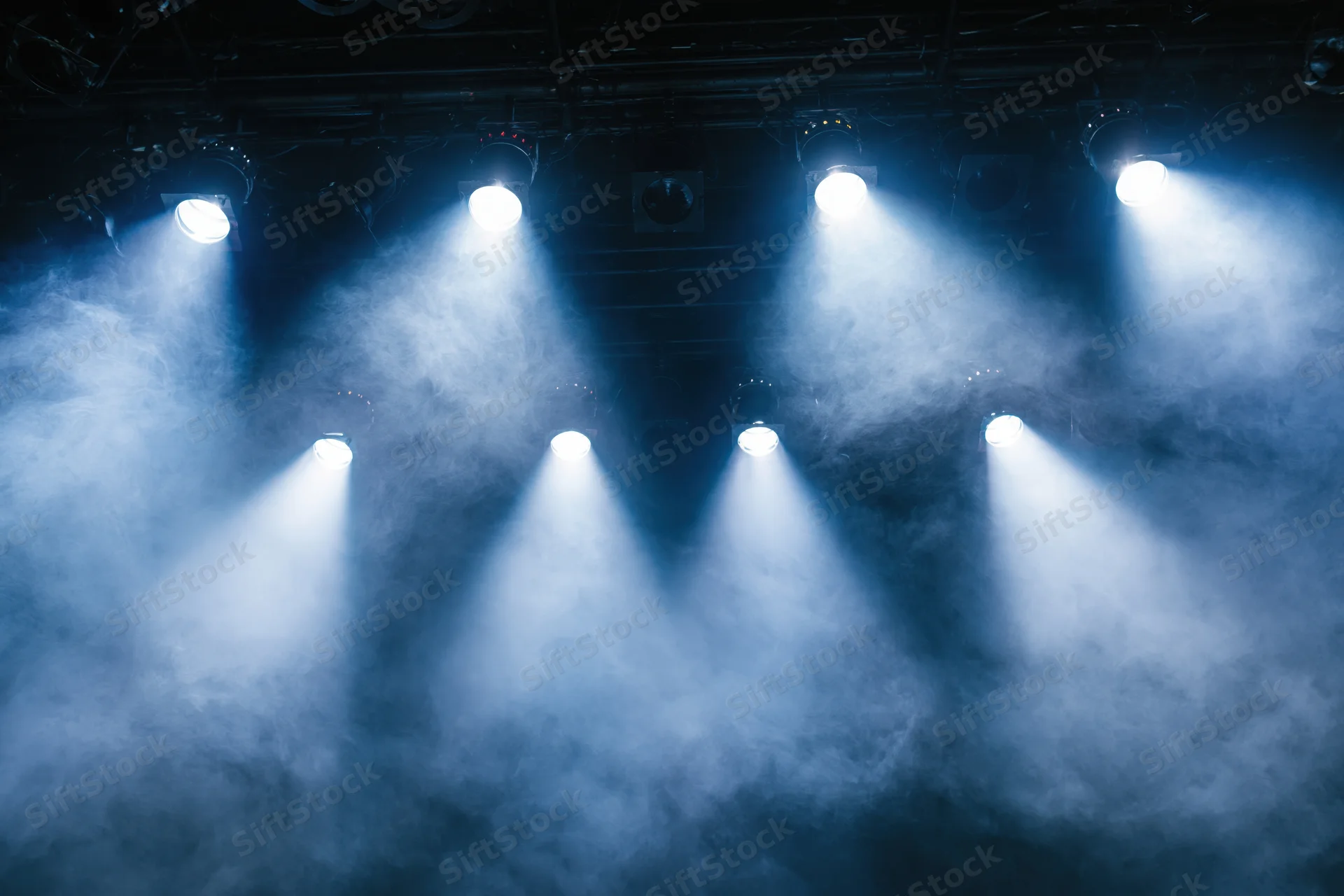 Multiple stage spotlights casting bright white and blue beams through smoke in a theater setting