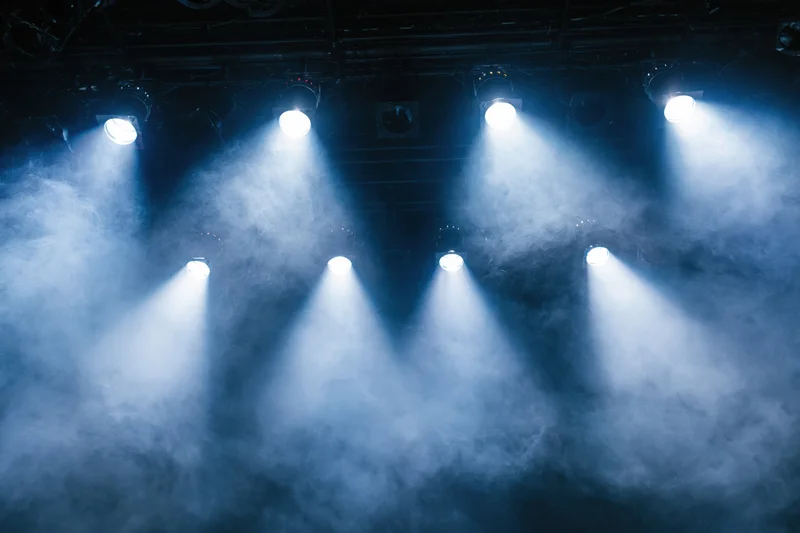 Multiple stage spotlights casting bright white and blue beams through smoke in a theater setting