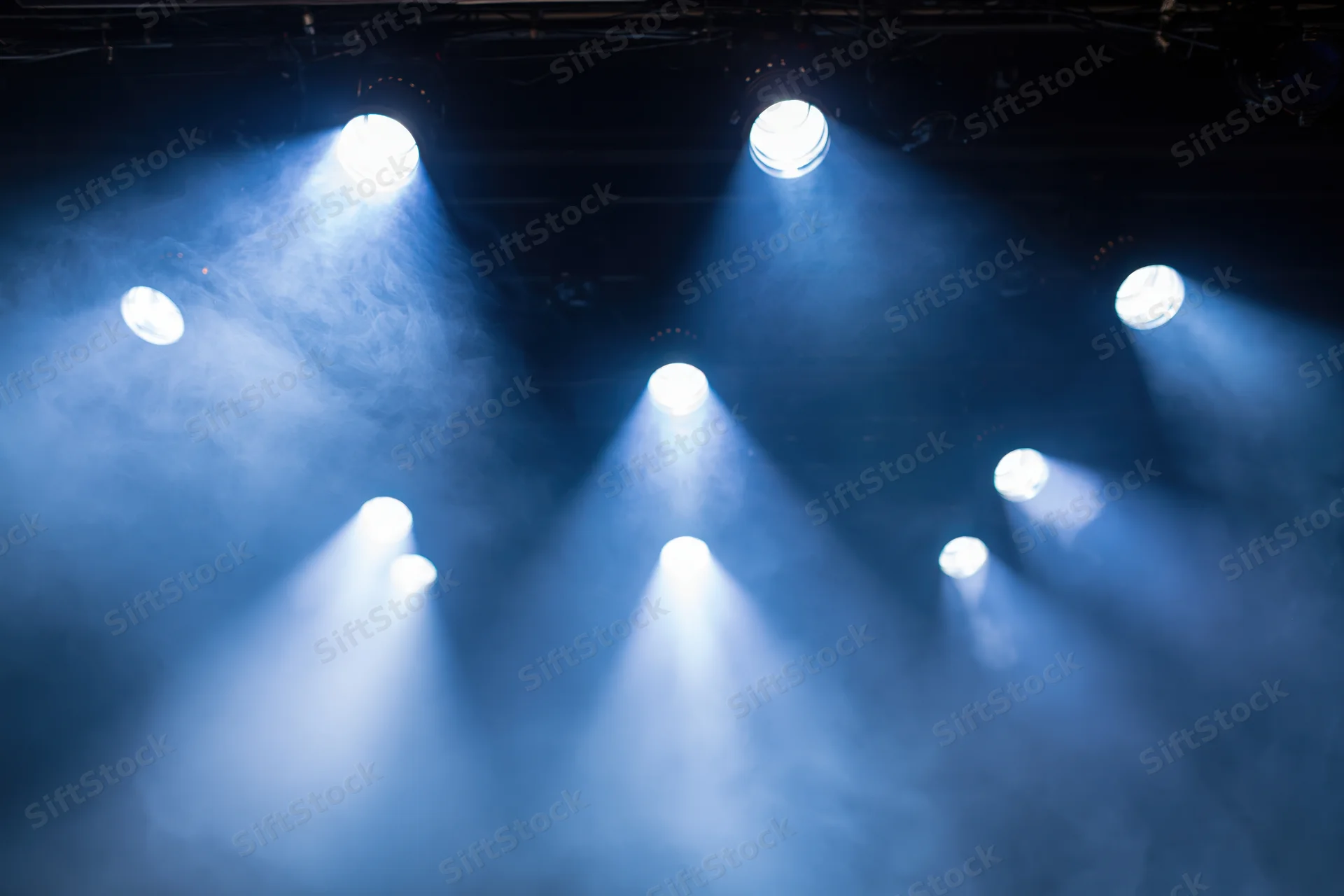 Multiple stage spotlights casting bright white and blue beams through smoke in a theater setting