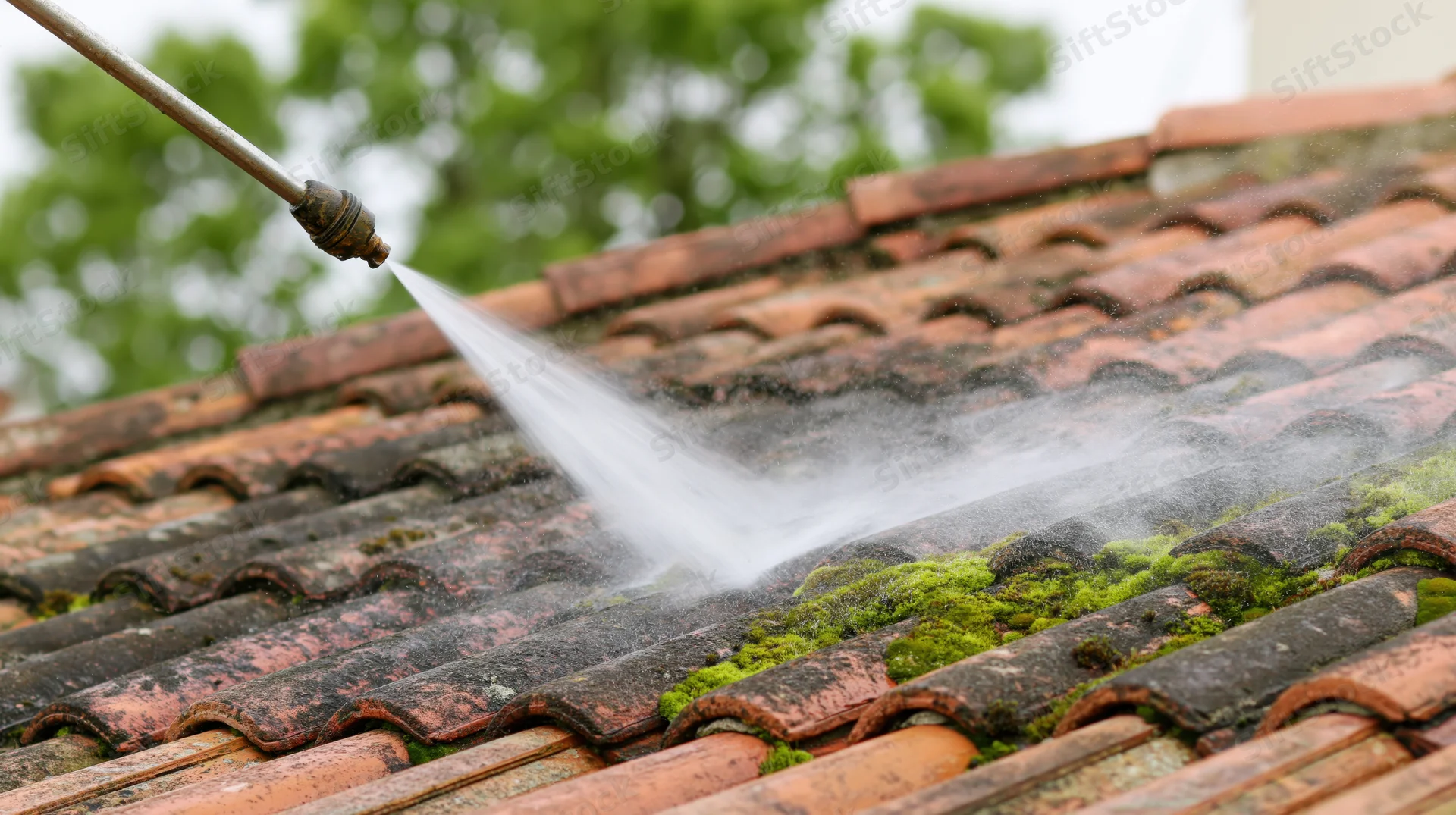 Pressure washer nozzle spraying water across moss-covered terracotta roof tiles during cleaning