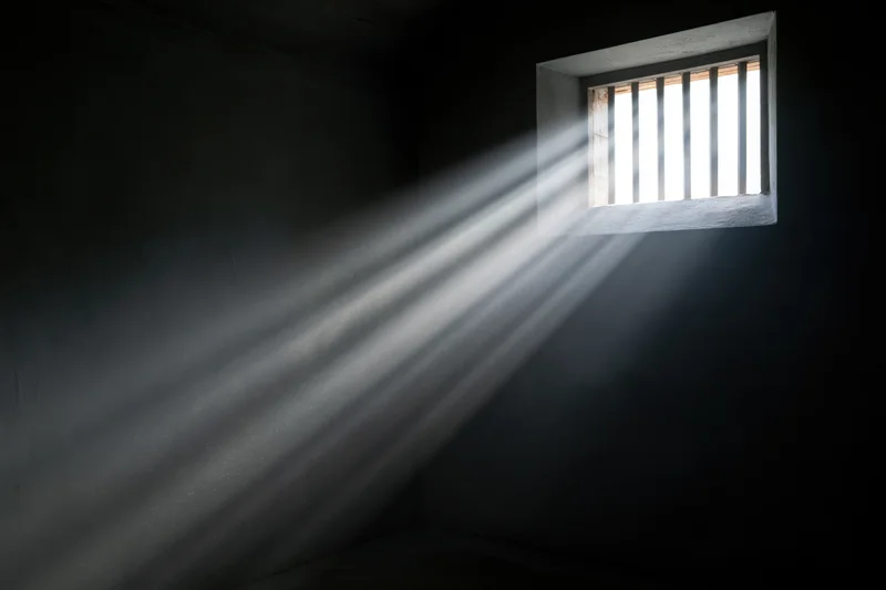 Rays of daylight streaming through a small barred window in a dark prison cell