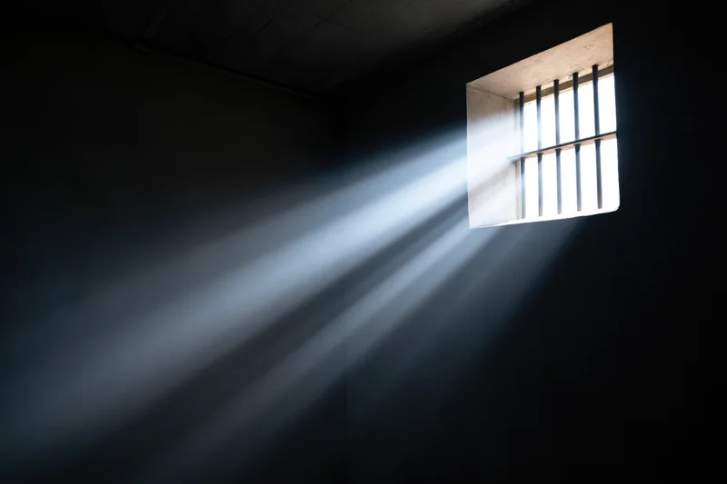 Rays of daylight streaming through a small barred window in a dark prison cell