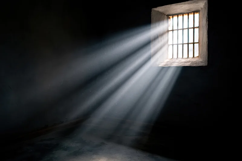 Rays of daylight streaming through a small barred window in a prison cell interior