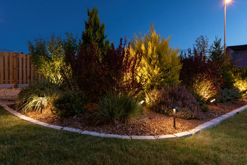Residential garden bed illuminated by low-voltage uplights at dusk with shrubs and mulch bordered by stone edging