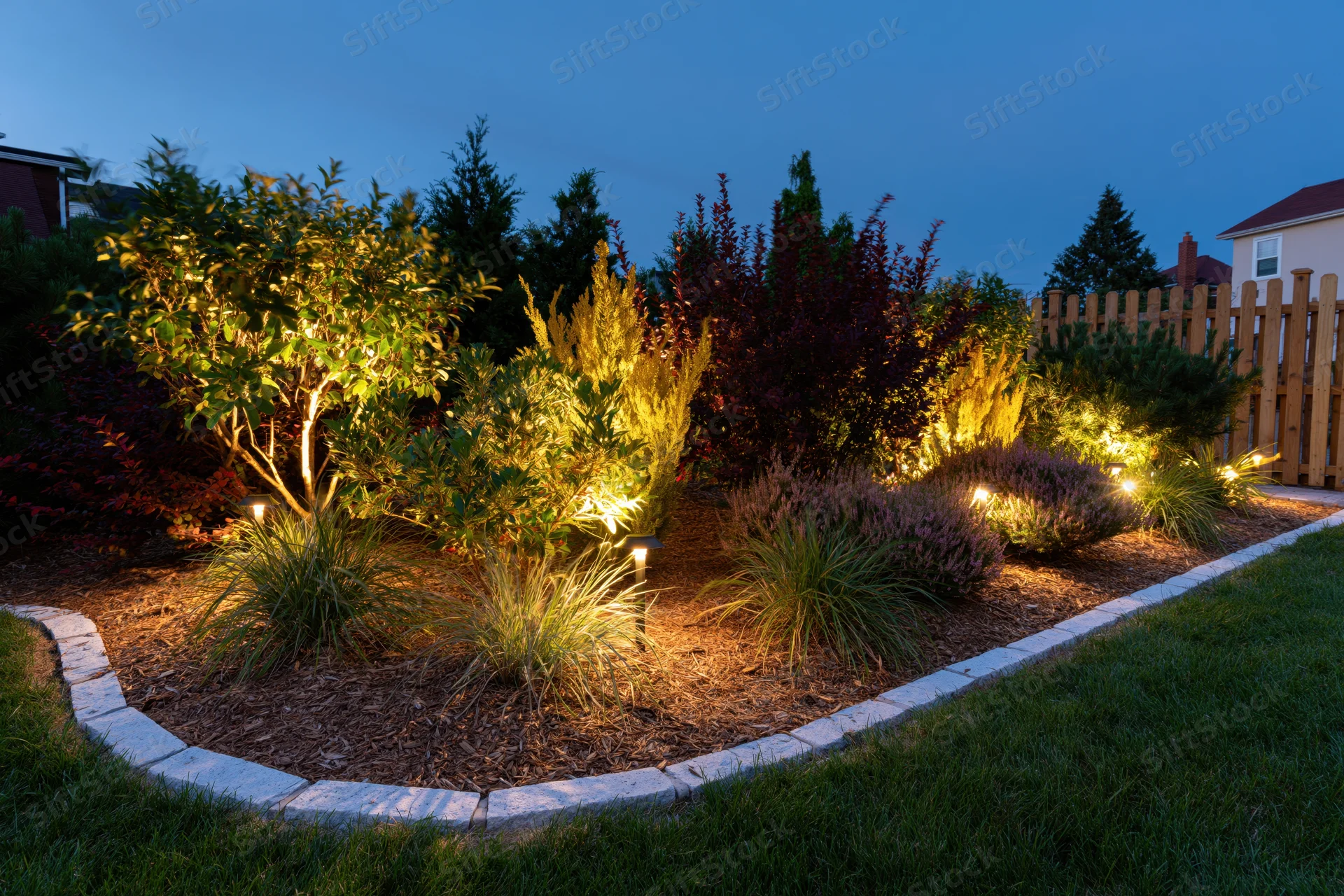 Residential garden bed illuminated by low-voltage uplighting at dusk with mulch, shrubs and ornamental grasses bordered by stone edging