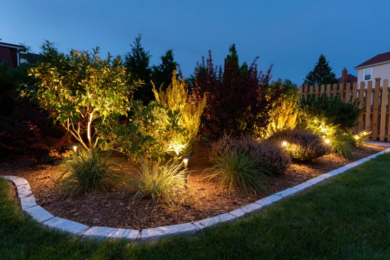 Residential garden bed illuminated by low-voltage uplighting at dusk with mulch, shrubs and ornamental grasses bordered by stone edging