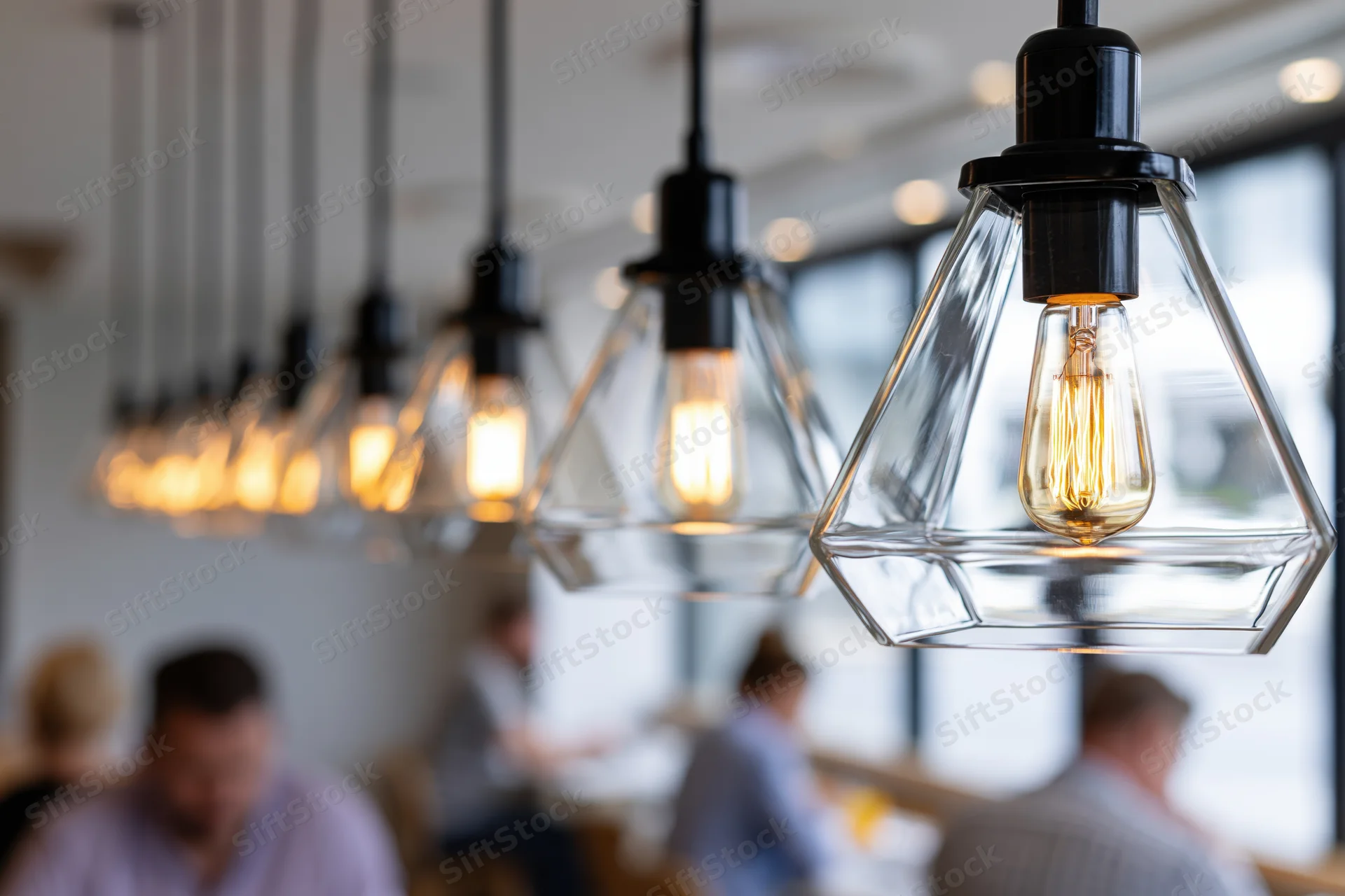 Row of geometric pyramid-shaped clear glass pendant lights with filament bulbs hanging over a dining area in a modern cafe