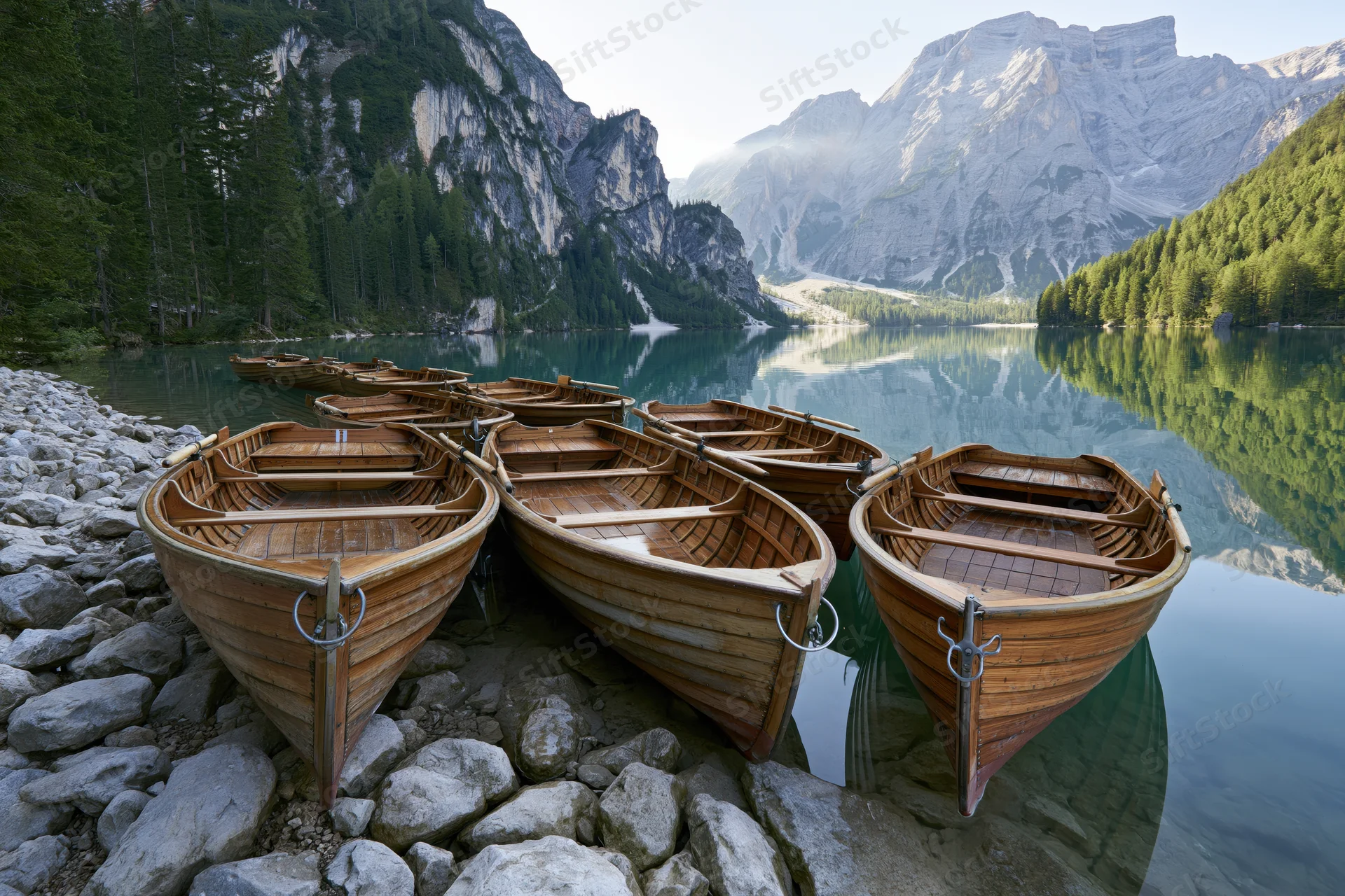 Row of wooden rowboats moored along rocky lakeshore with mountain reflection in calm water