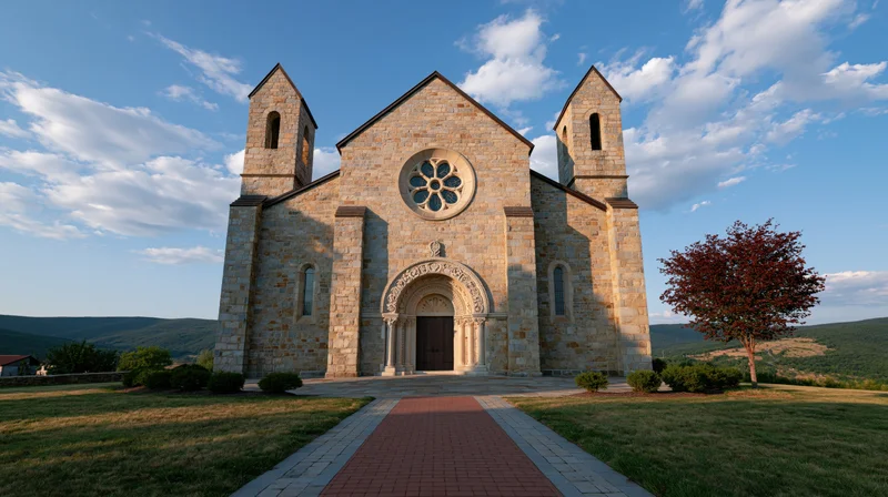 Small roman stone church with rose window and twin bell towers on a grassy hill under a blue sky with scattered clouds