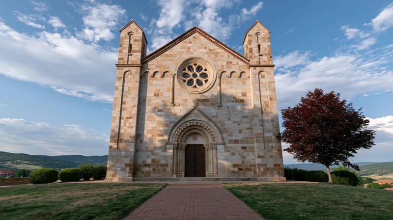 Small roman stone church with rose window and ornate arched entrance on a grassy hill under blue sky with clouds