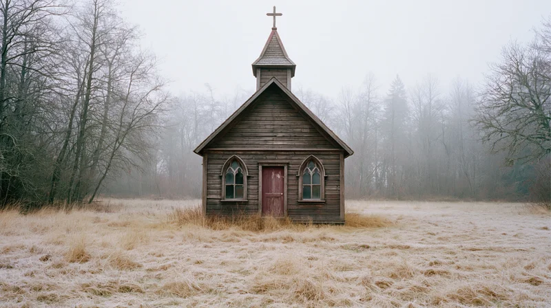 Small abandoned wooden chapel with cross-topped steeple in frosty field surrounded by bare trees