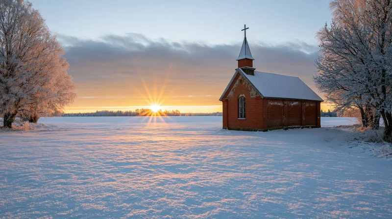 Small red brick chapel with cross-topped steeple standing in snowy field at sunrise