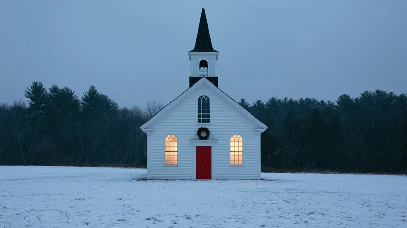 Small white clapboard church with black steeple and red door in snowy field at dusk