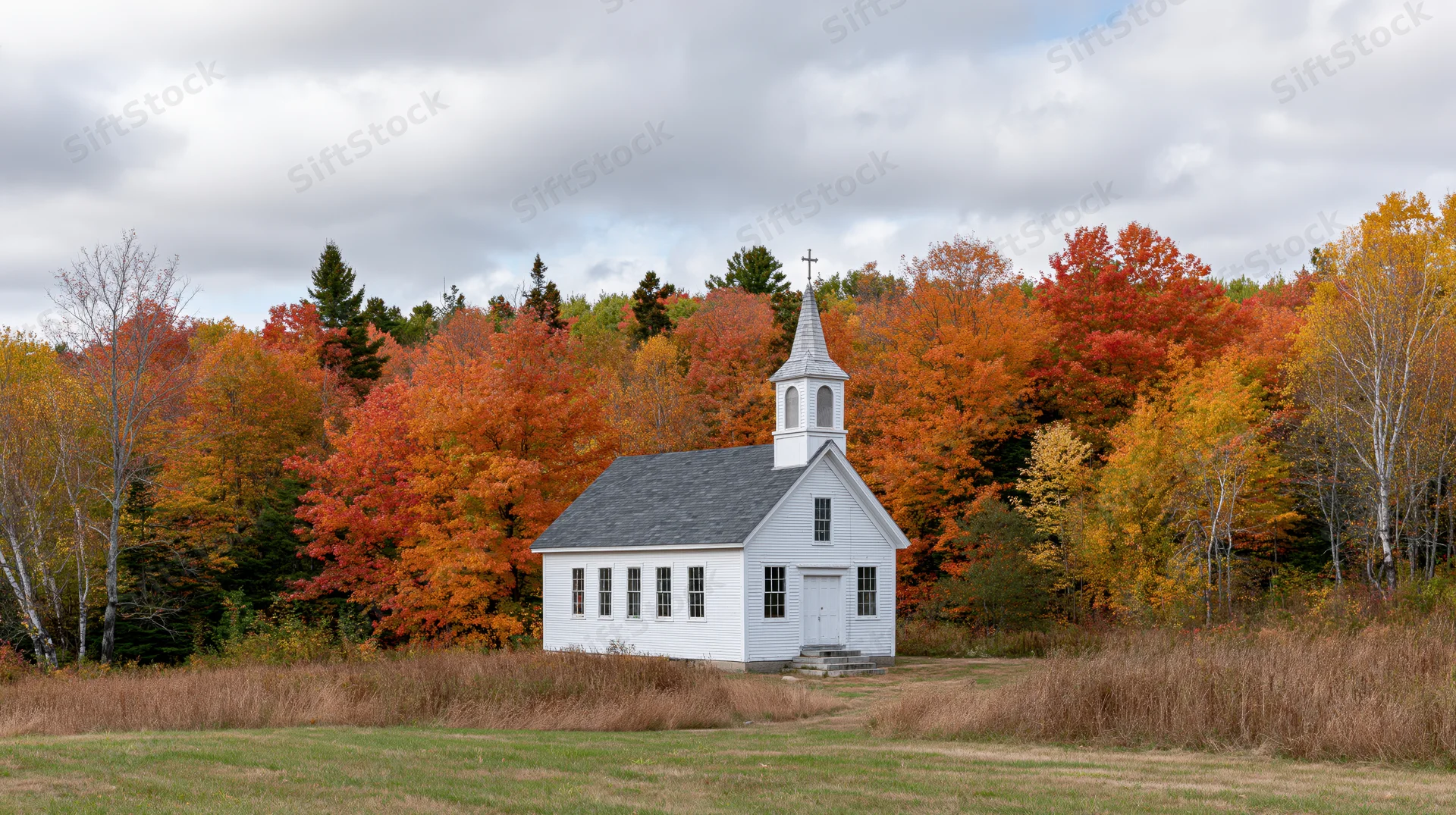 Small white clapboard church with steeple and cross surrounded by autumn foliage in rural new england landscape