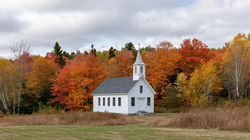 Small white clapboard church with steeple and cross surrounded by autumn foliage in rural new england landscape