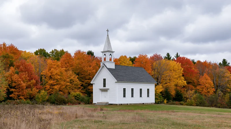 Small white clapboard church with steeple and cross in a grassy field surrounded by autumn foliage under a cloudy sky