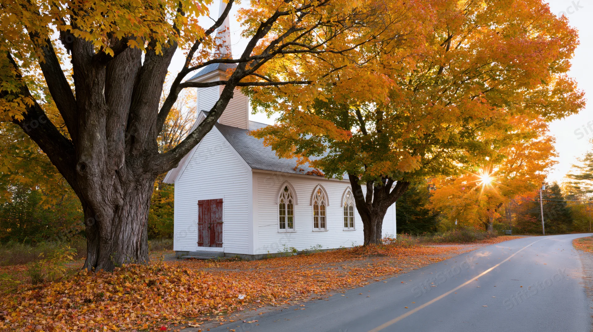 Small white clapboard church with steeple beside autumn trees and rural road