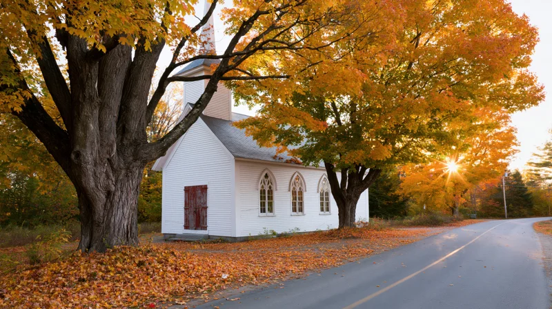 Small white clapboard church with steeple beside autumn trees and rural road
