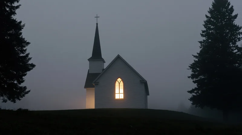 Small white clapboard church with illuminated stained glass window on foggy hill at twilight