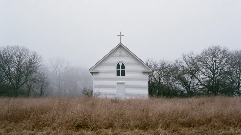 Small white clapboard church with rooftop cross in a foggy rural field surrounded by bare trees and dry grass