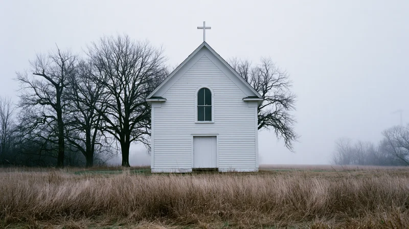 Small white clapboard church with rooftop cross in a foggy rural field