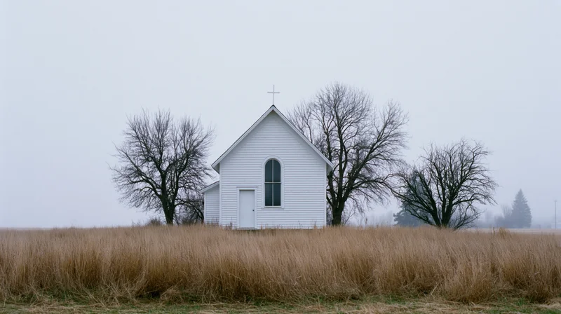 Small white clapboard church with rooftop cross in a dry grassy field surrounded by bare trees under an overcast sky
