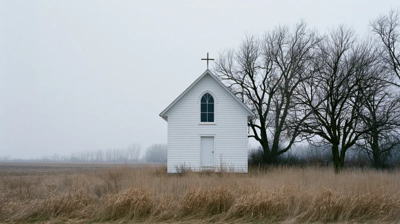 Small white clapboard church with rooftop cross standing in a dry grassy field beside bare trees under an overcast sky
