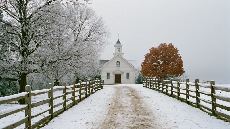 Small white clapboard church with steeple at end of snow-covered path lined with wooden fence in rural winter landscape