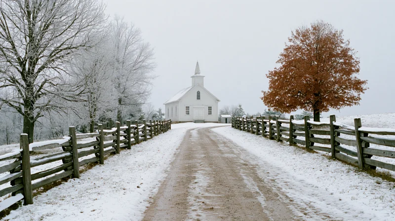 Small white clapboard church with steeple at end of snow-covered dirt road lined with wooden fence and bare trees in rural winter landscape