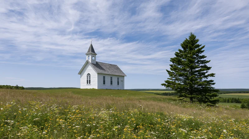 Small white clapboard church with steeple and cross on a grassy hill in a rural field under a blue sky with scattered clouds