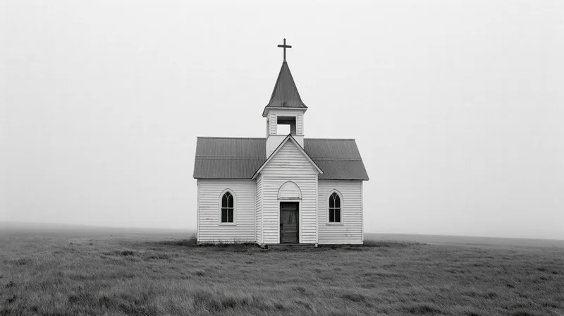 Small white clapboard church with steeple and cross standing alone in a grassy prairie field under an overcast sky