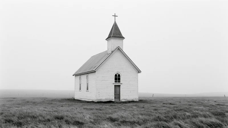 Small white clapboard church with steeple and cross standing alone in a grassy prairie field under overcast sky