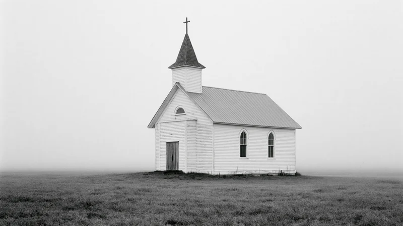 Small white clapboard church with steeple and cross standing alone in a foggy prairie field