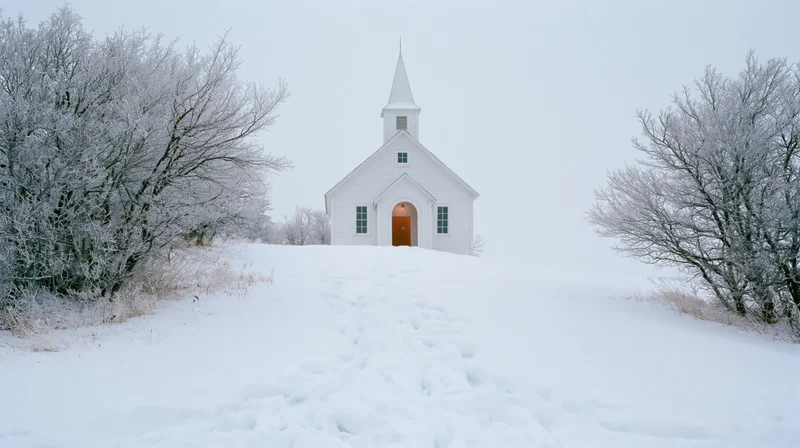 Small white country church with pointed steeple standing on snowy hill, surrounded by frosted trees and footprints in snow