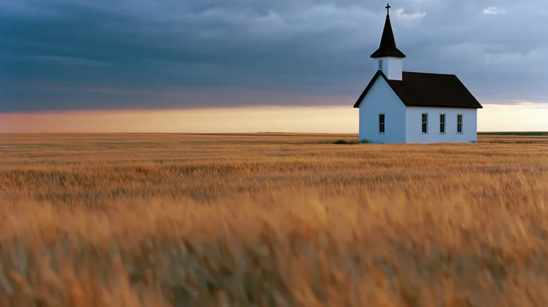 Small white chapel with dark steeple and cross standing in a golden wheat field at dusk