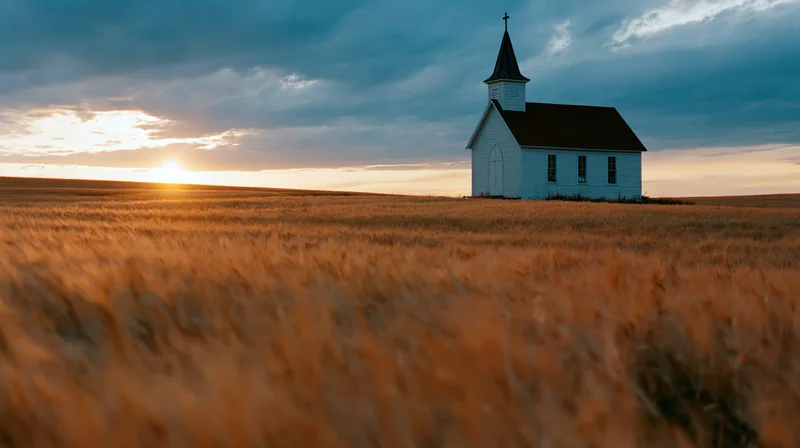 Small white chapel with dark steeple and cross standing in a golden wheat field at sunset