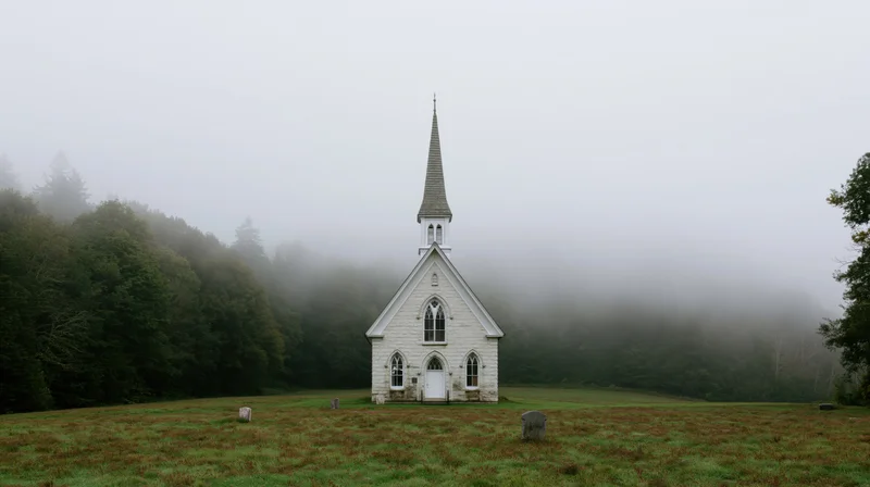 Small white stone gothic revival church with tall spire standing in a foggy field surrounded by forest and tombstones