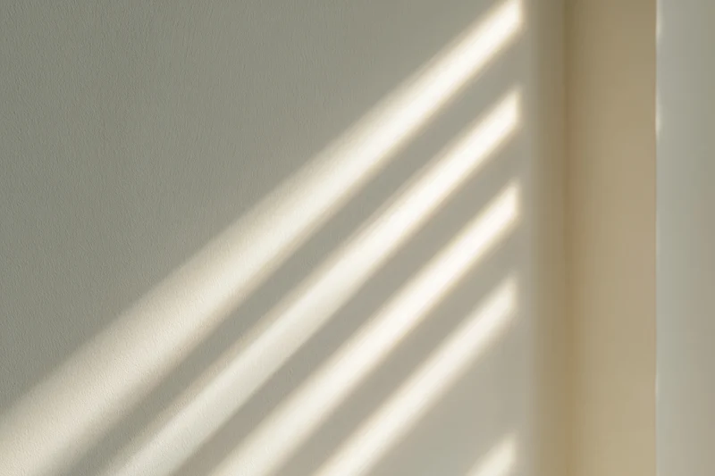 Diagonal sunlight shadows cast across a plain off-white interior wall corner