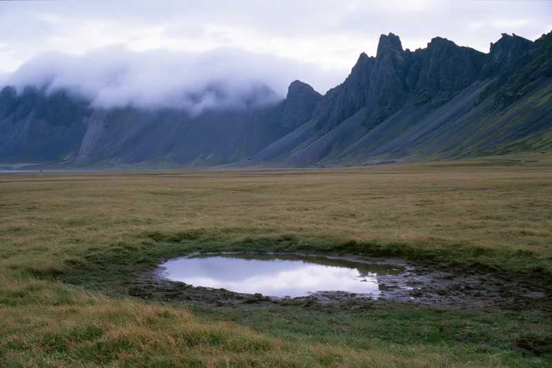Flat grassland with a small puddle reflecting cloudy sky, bordered by jagged dark volcanic mountains in the distance