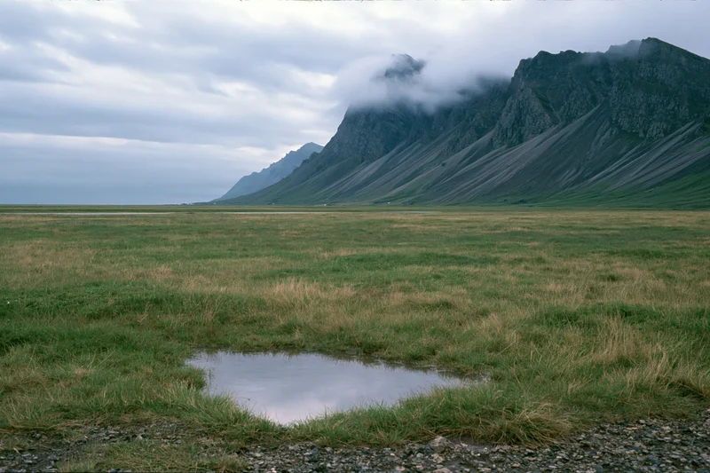 Vast flat grassland stretching toward jagged dark volcanic mountains under cloudy sky