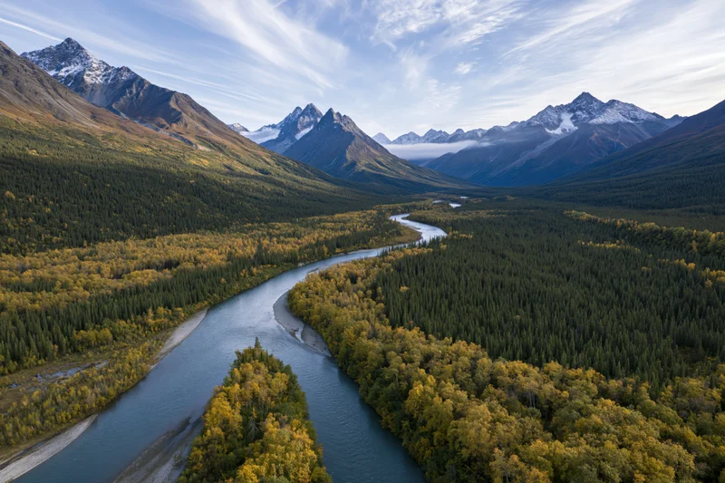 Winding river cutting through alpine valley surrounded by snow-capped mountains and autumn forest