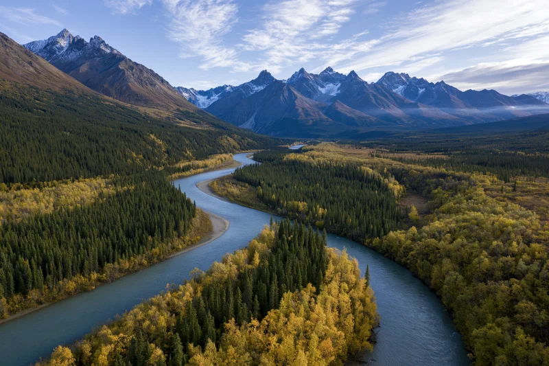 Winding river cutting through alpine valley surrounded by snow-capped mountains and autumn forest