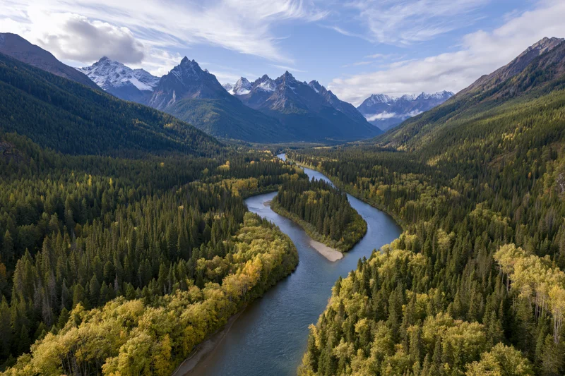 Winding river cutting through alpine valley surrounded by snow-capped mountains and dense evergreen forest in autumn