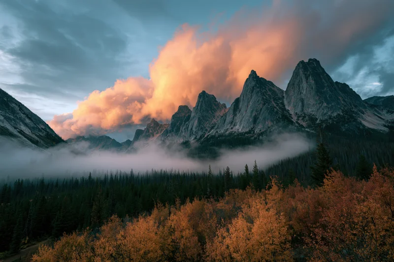 Jagged rocky mountain peaks rising above foggy valley with autumn trees in foreground at twilight