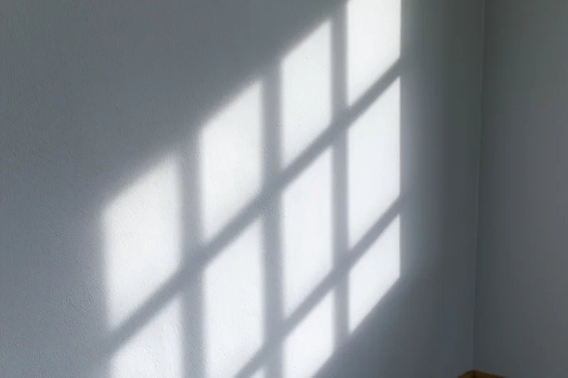 Diagonal window grid shadow cast across plain light gray wall in interior space