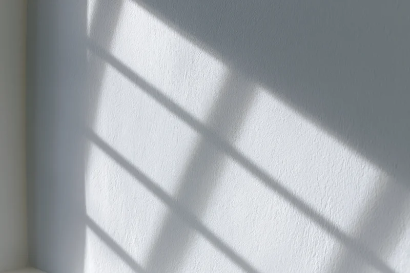 Diagonal window grid shadow cast across white textured wall in interior corner