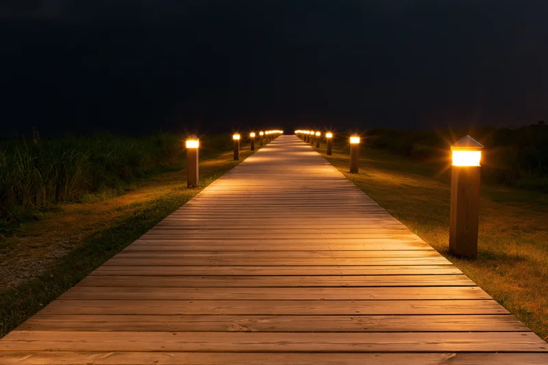 Wooden plank walkway illuminated by warm lights at night extending through grassy dunes toward darkness